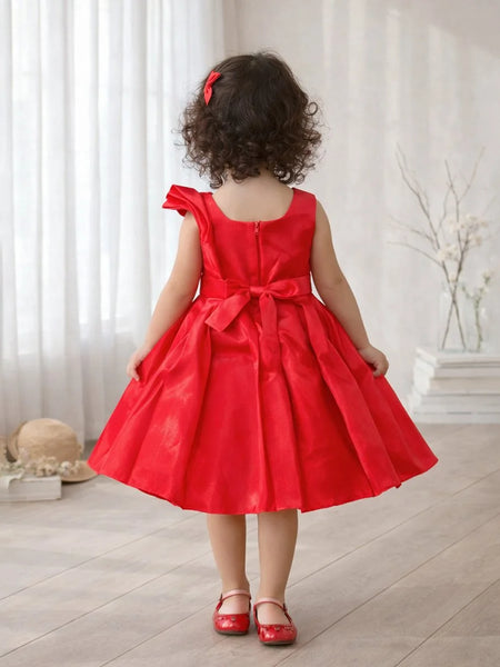 Child wearing a red dress with a bow in a room with white curtains and a vase.