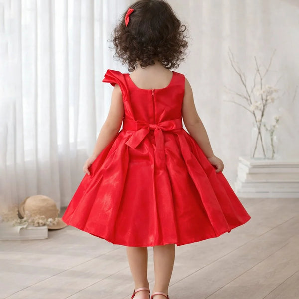 Child wearing a red dress with a bow in a room with white curtains and a vase.
