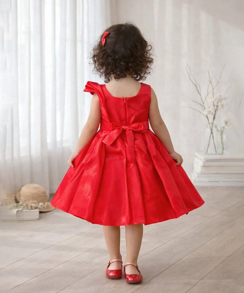 Child wearing a red dress with a bow in a room with white curtains and a vase.