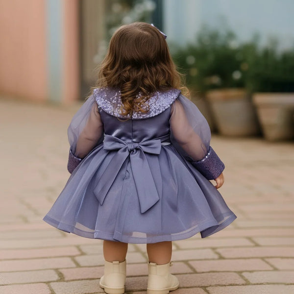 Child wearing a purple dress with a large bow, standing on a paved walkway.