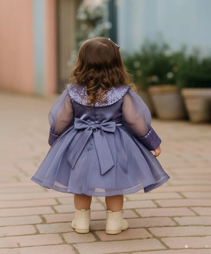 Child wearing a purple dress with a large bow, standing on a paved walkway.