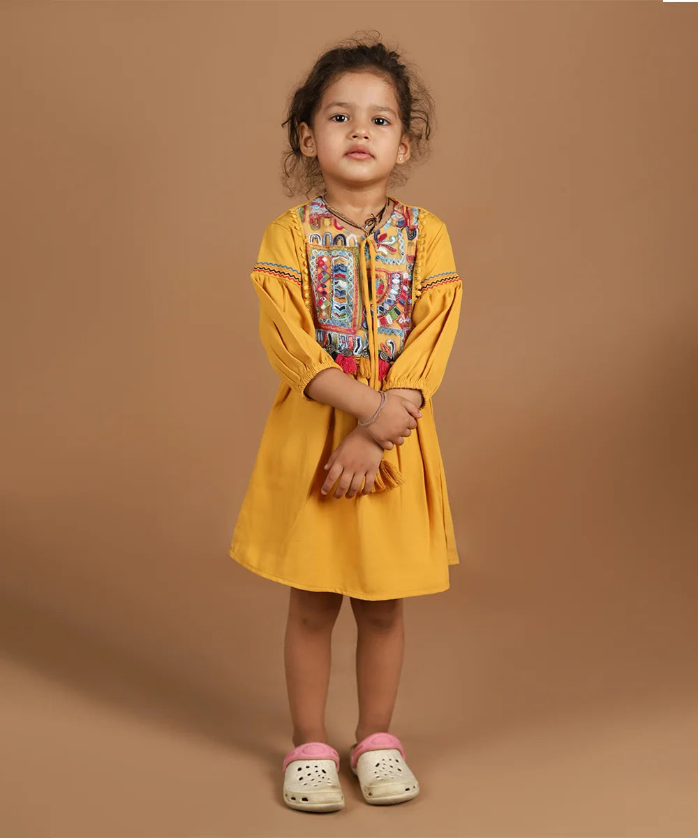 It’s a mustard color dress that comes with net embroidered fabric and tassel detailing on the dress. It features a laced outline around the embroidered net fabric and tie knot strings on the dress.