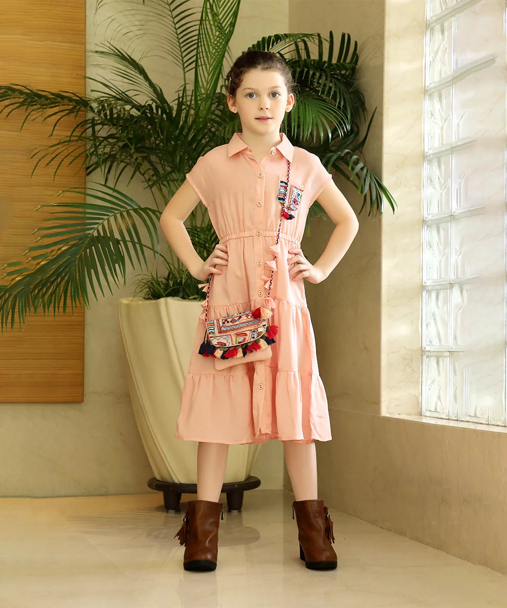 It is a peach Colored tiered shirt dress for girls that comes with a matching sling bag. It features a pocket, an elasticated waist and embroidery detailing on pocket as well as on the bag.