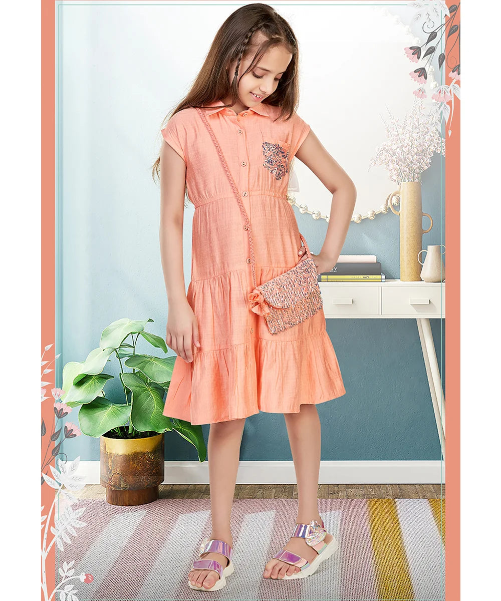  It’s a peach Colored tiered trendy dresses for kids that comes with a matching sling bag for party looks for girls. It features an elasticated waist, pocket and sequin detailing on the pocket as well as on the bag.