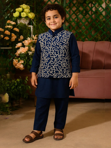 Child wearing a navy blue traditional outfit with a patterned vest, standing in front of flowers and a pink couch.