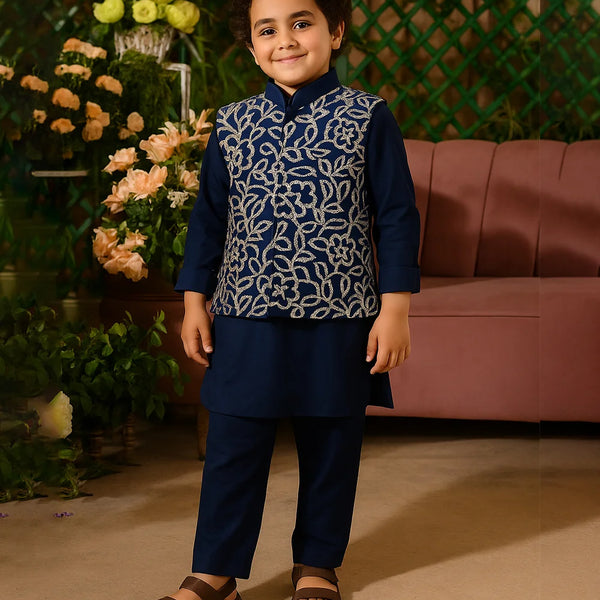 Child wearing a navy blue traditional outfit with a patterned vest, standing in front of flowers and a pink couch.