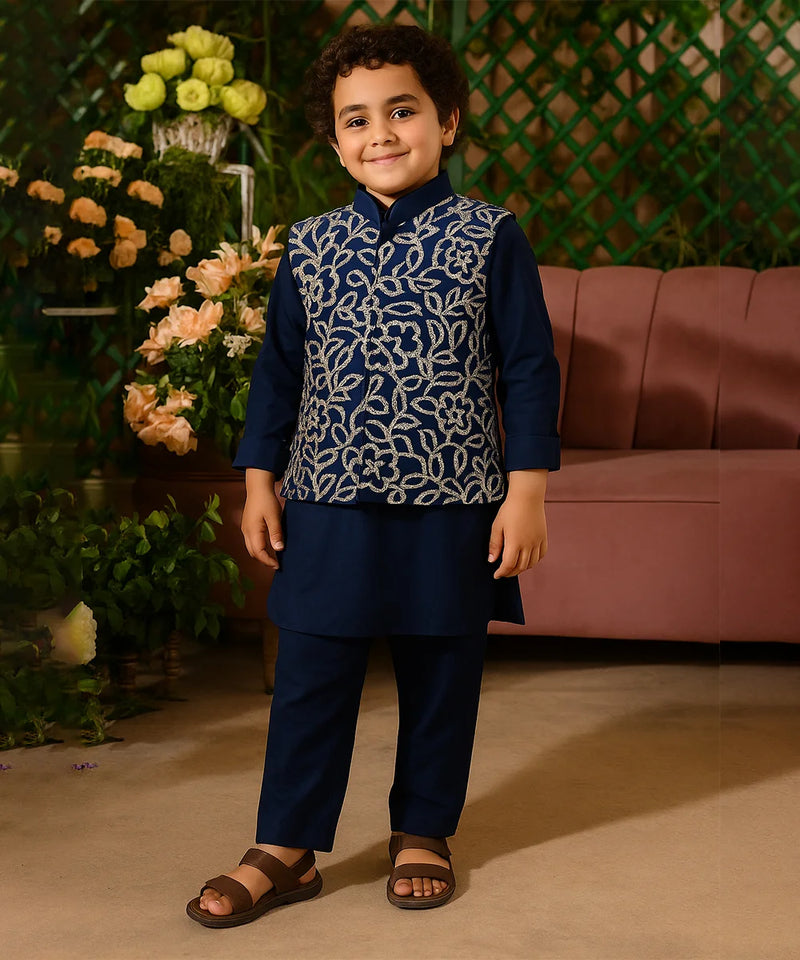 Child wearing a navy blue traditional outfit with a patterned vest, standing in front of flowers and a pink couch.