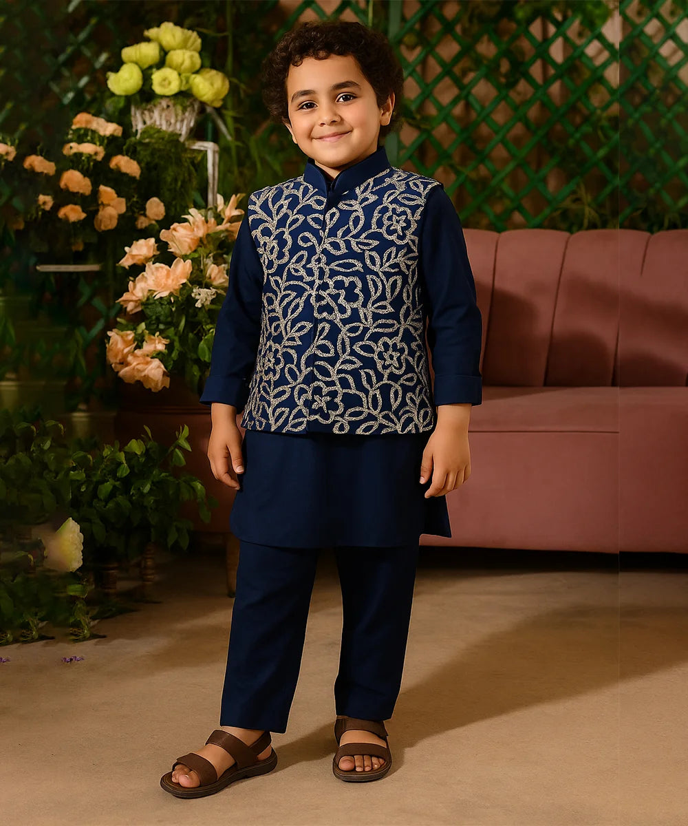 Child wearing a navy blue traditional outfit with a patterned vest, standing in front of flowers and a pink couch.