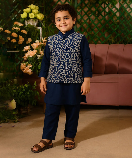 Child wearing a navy blue traditional outfit with a patterned vest, standing in front of flowers and a pink couch.