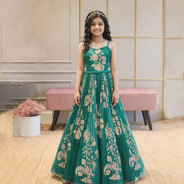 Young girl in a teal embroidered dress standing indoors with a pink bench and decorative plants in the background.
