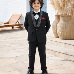 Young boy in a formal black suit with a bow tie standing outdoors.