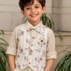 Young boy wearing a floral vest and bow tie, standing outdoors with plants in the background.
