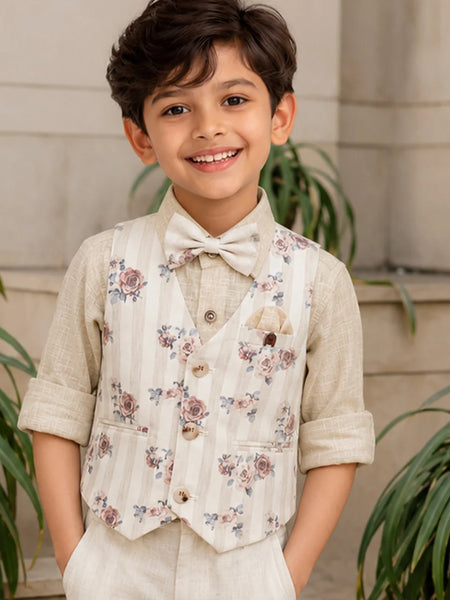 Young boy wearing a floral vest and bow tie, standing outdoors with plants in the background.