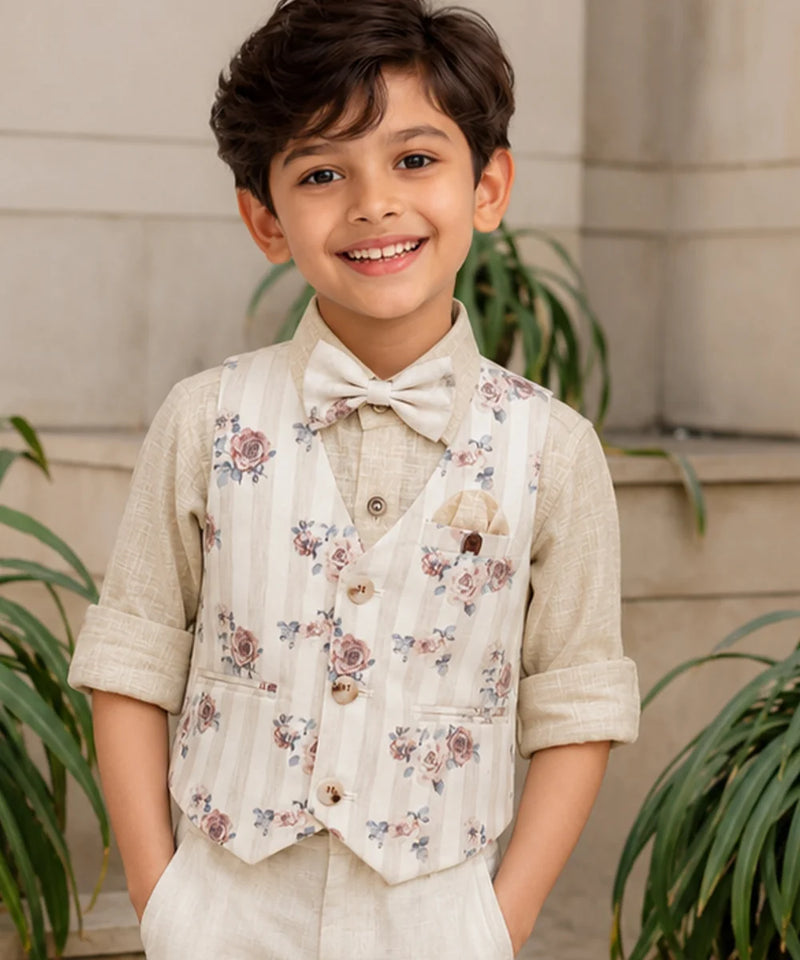 Young boy wearing a floral vest and bow tie, standing outdoors with plants in the background.