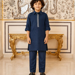 A young boy modeling a navy blue kurta and trouser set with a decorative silver-gray embroidered placket, standing in front of an ornate gold console table and a patterned wall.