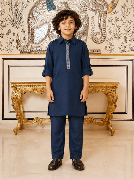 A young boy modeling a navy blue kurta and trouser set with a decorative silver-gray embroidered placket, standing in front of an ornate gold console table and a patterned wall.