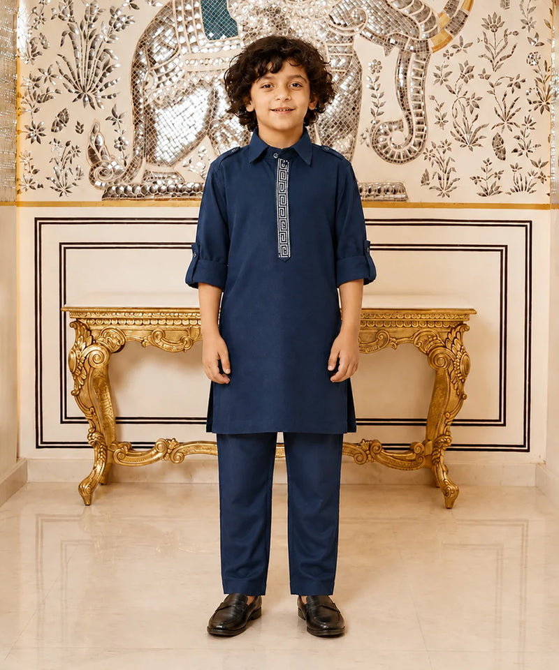 A young boy modeling a navy blue kurta and trouser set with a decorative silver-gray embroidered placket, standing in front of an ornate gold console table and a patterned wall.