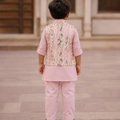 A young boy viewed from behind wearing a light pink traditional outfit with a patterned vest and matching trousers, standing in an outdoor courtyard.
