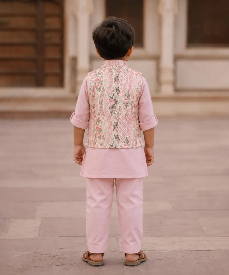 A young boy viewed from behind wearing a light pink traditional outfit with a patterned vest and matching trousers, standing in an outdoor courtyard.