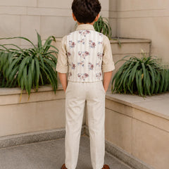 Child wearing a floral vest and beige pants standing in front of a stone wall with plants.