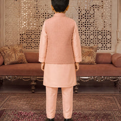 A young boy viewed from behind wearing a peach-colored traditional Indian outfit consisting of a kurta and trousers, standing in front of an ornate carved wooden screen and sofa.