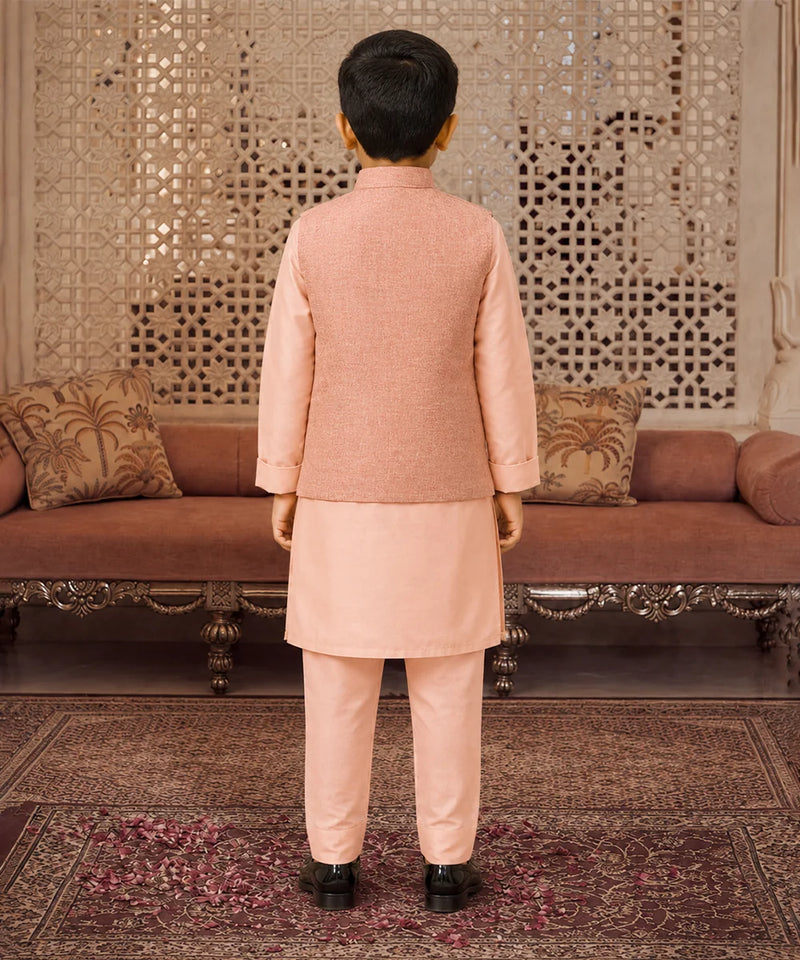 A young boy viewed from behind wearing a peach-colored traditional Indian outfit consisting of a kurta and trousers, standing in front of an ornate carved wooden screen and sofa.