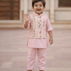 A young child wearing a pink traditional outfit with a floral embroidered vest and matching pants, standing outdoors and waving.