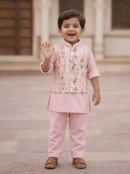A young child wearing a pink traditional outfit with a floral embroidered vest and matching pants, standing outdoors and waving.