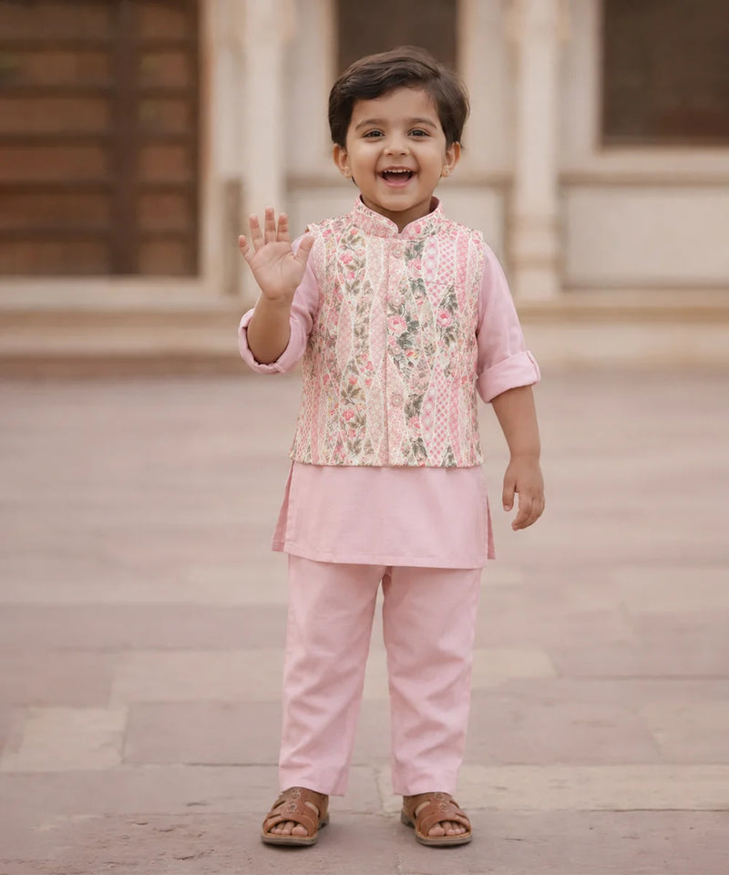 A young child wearing a pink traditional outfit with a floral embroidered vest and matching pants, standing outdoors and waving.
