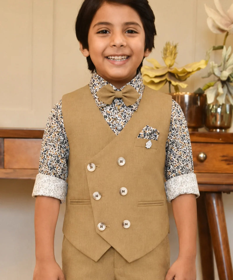 Child wearing a formal outfit with a vest and bow tie, standing in front of a wooden table with decorative items.