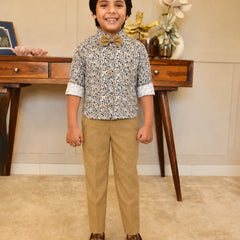 Child wearing a patterned shirt and beige pants standing in a room with wooden furniture and decorative items.
