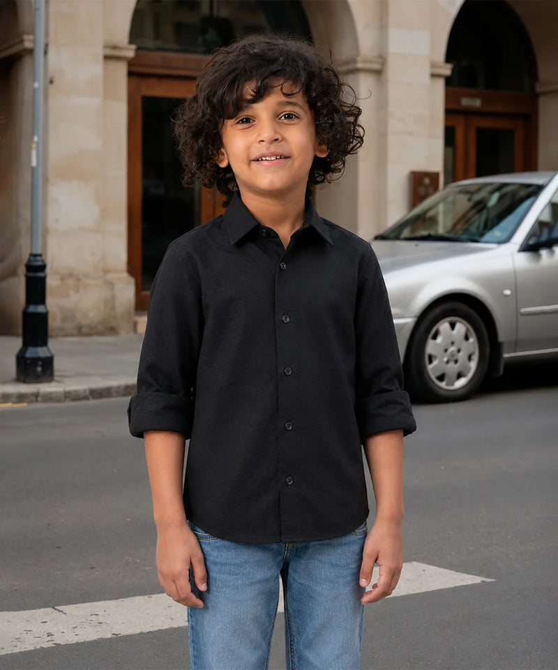 Child wearing a black shirt and blue jeans standing on a street with a car and building in the background.