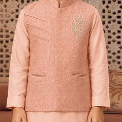 A child wearing a peach-pink traditional Indian outfit with a textured jacket featuring diagonal welt pockets and a floral embroidery on the chest, paired with a matching skirt. The background shows a decorative wooden screen.