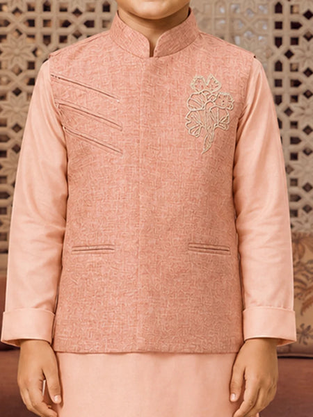 A child wearing a peach-pink traditional Indian outfit with a textured jacket featuring diagonal welt pockets and a floral embroidery on the chest, paired with a matching skirt. The background shows a decorative wooden screen.