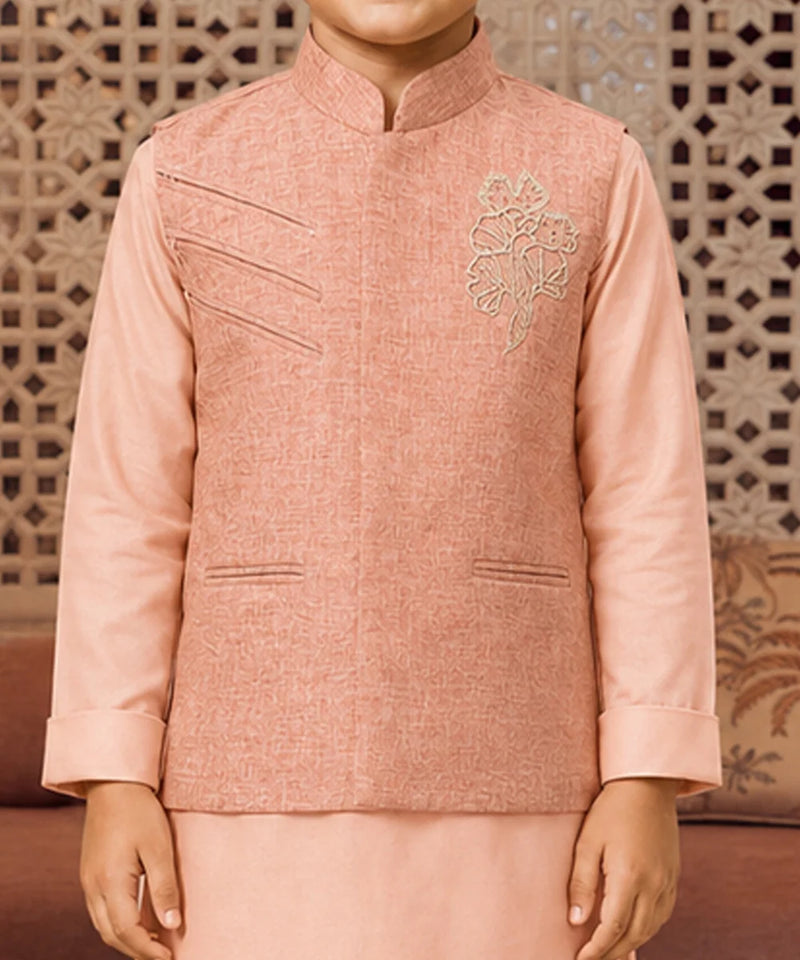 A child wearing a peach-pink traditional Indian outfit with a textured jacket featuring diagonal welt pockets and a floral embroidery on the chest, paired with a matching skirt. The background shows a decorative wooden screen.