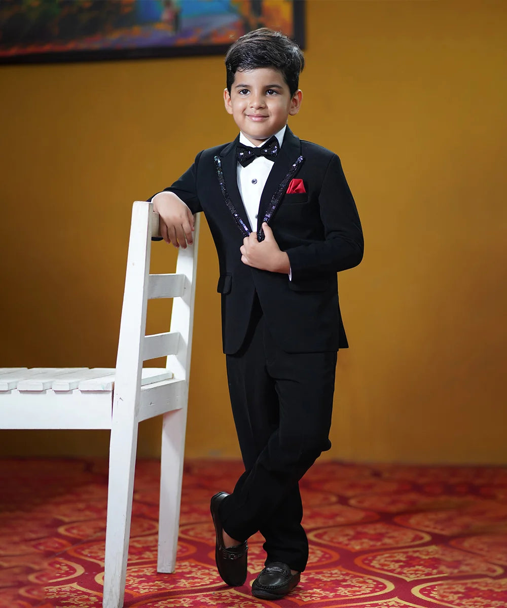 This classic tuxedo is rendered in black color and comes with matching pants, a white shirt, a black bow and a red pocket square. It features a beautiful sequin detailing on the collar and a bow curated from a black sequin fabric.