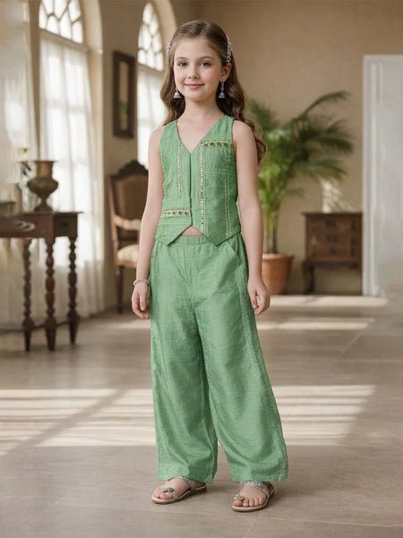 Young girl in a green outfit standing in a well-lit room with wooden floors and large windows.