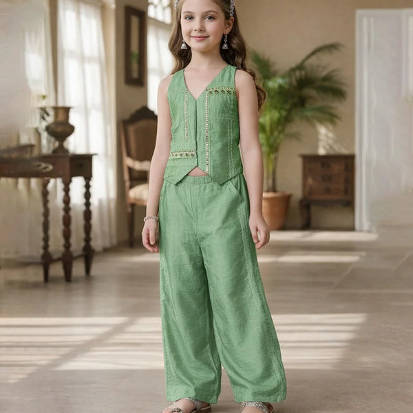Young girl in a green outfit standing in a well-lit room with wooden floors and large windows.