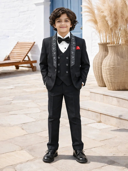 Young boy in a formal black suit with a bow tie standing outdoors.