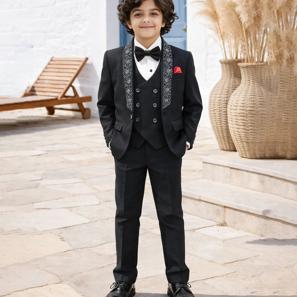 Young boy in a formal black suit with a bow tie standing outdoors.