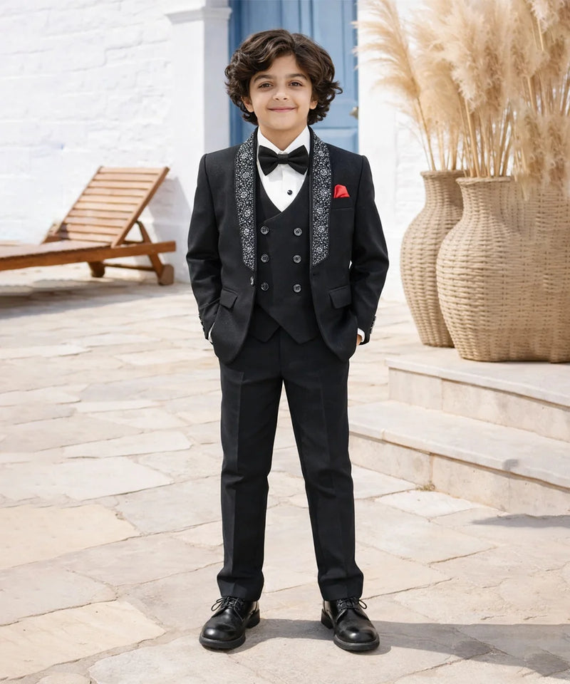 Young boy in a formal black suit with a bow tie standing outdoors.
