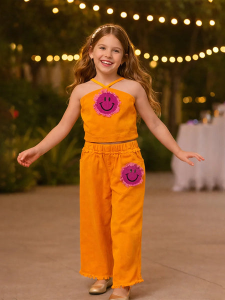 Young girl in an orange outfit with smiley face designs, standing outdoors with string lights in the background.