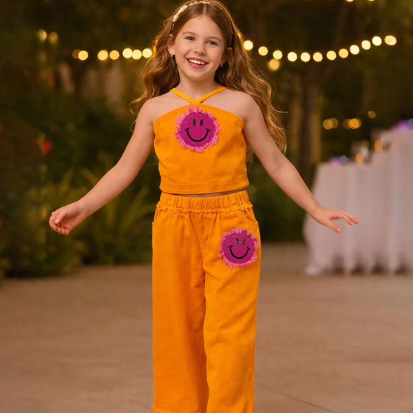 Young girl in an orange outfit with smiley face designs, standing outdoors with string lights in the background.