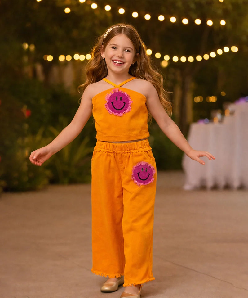 Young girl in an orange outfit with smiley face designs, standing outdoors with string lights in the background.