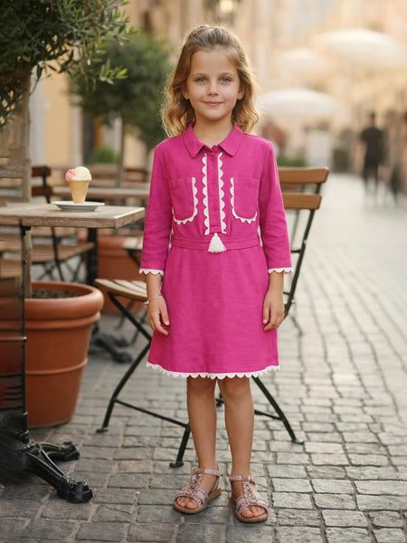 Young girl in a pink dress standing on a street with outdoor cafe furniture in the background.