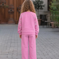 Person wearing a pink outfit standing on a paved area with a wooden door and plants in the background.