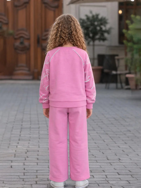 Person wearing a pink outfit standing on a paved area with a wooden door and plants in the background.