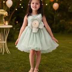 Young girl in a light green dress with a large flower design standing outdoors with balloons and lights in the background.