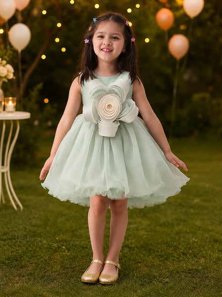 Young girl in a light green dress with a large flower design standing outdoors with balloons and lights in the background.