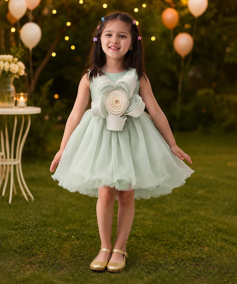 Young girl in a light green dress with a large flower design standing outdoors with balloons and lights in the background.